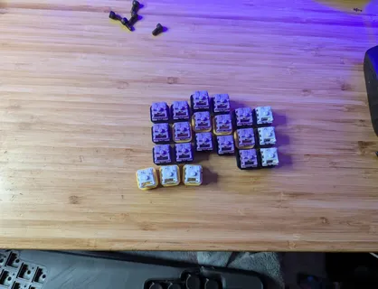 A collection of custom mechanical keyboard keycaps, including several sets of purple, white, and other colored keycaps, are arranged on a wooden desk surface