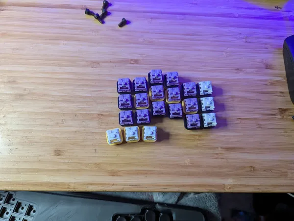 A collection of custom mechanical keyboard keycaps, including several sets of purple, white, and other colored keycaps, are arranged on a wooden desk surface