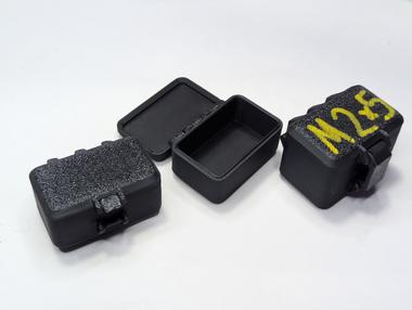 beginning of little boxes Four little 3d printed boxes laying on a white background, one contains a painted lid that says M 2x5.